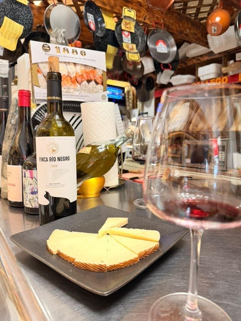 A glass of red beside a bottle and a plate of sliced cheese on a counter