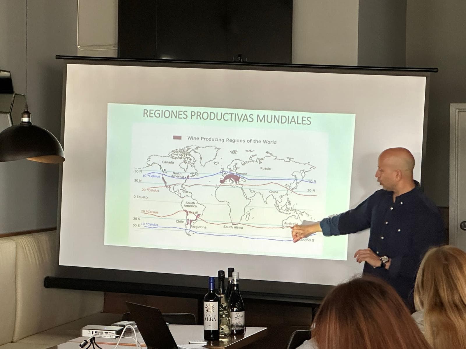An instructor pointing at a projected map of the wine regions of the world, bottles on the table in front