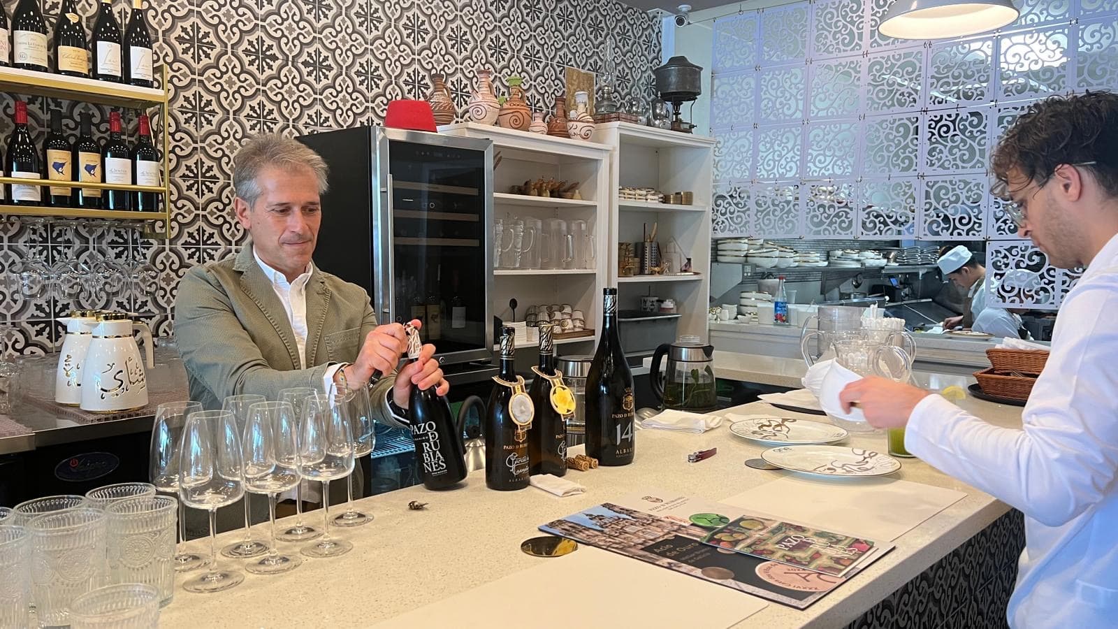 A sommelier pouring for a chef at a restaurant counter lined with glasses and open bottles