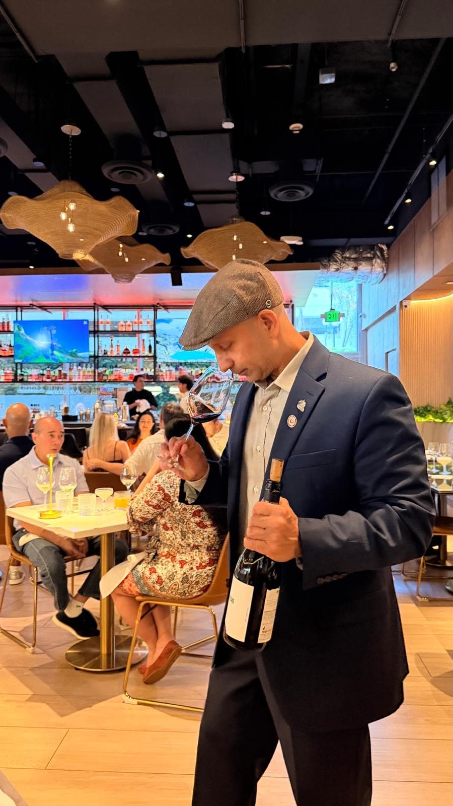 A sommelier in a flat cap nosing a glass while holding the bottle, guests seated behind